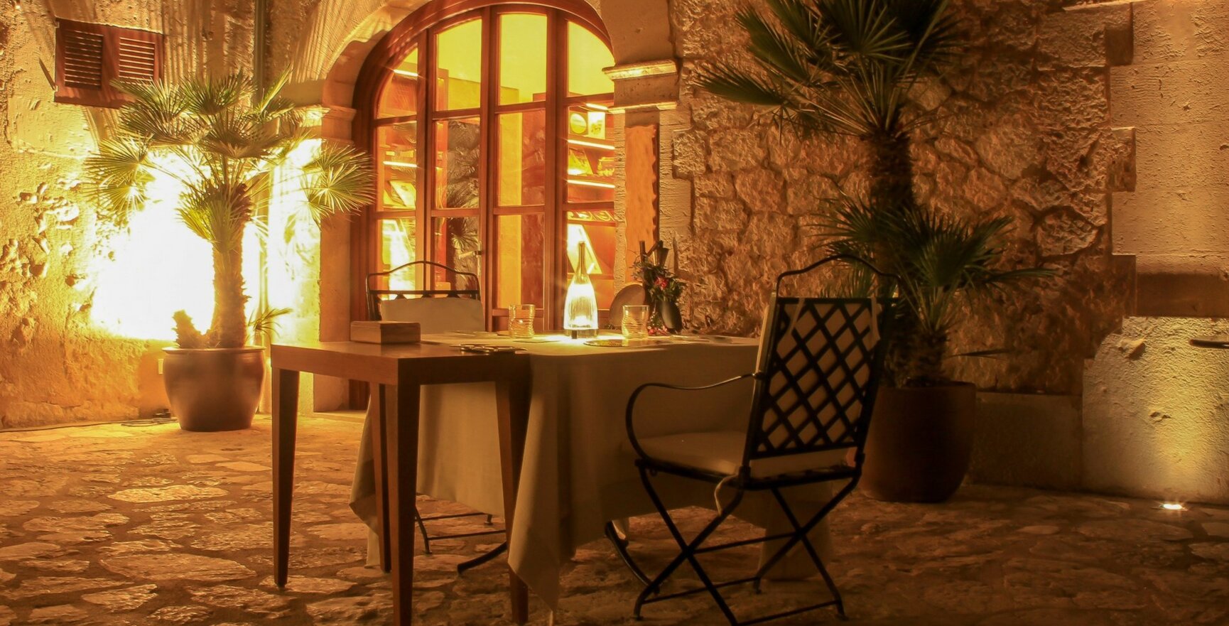 Intimer Außenbereich mit Steinhwänden, warmer Beleuchtung, Palmen in Töpfen und gedecktem Tisch mit Flasche und Gläsern
