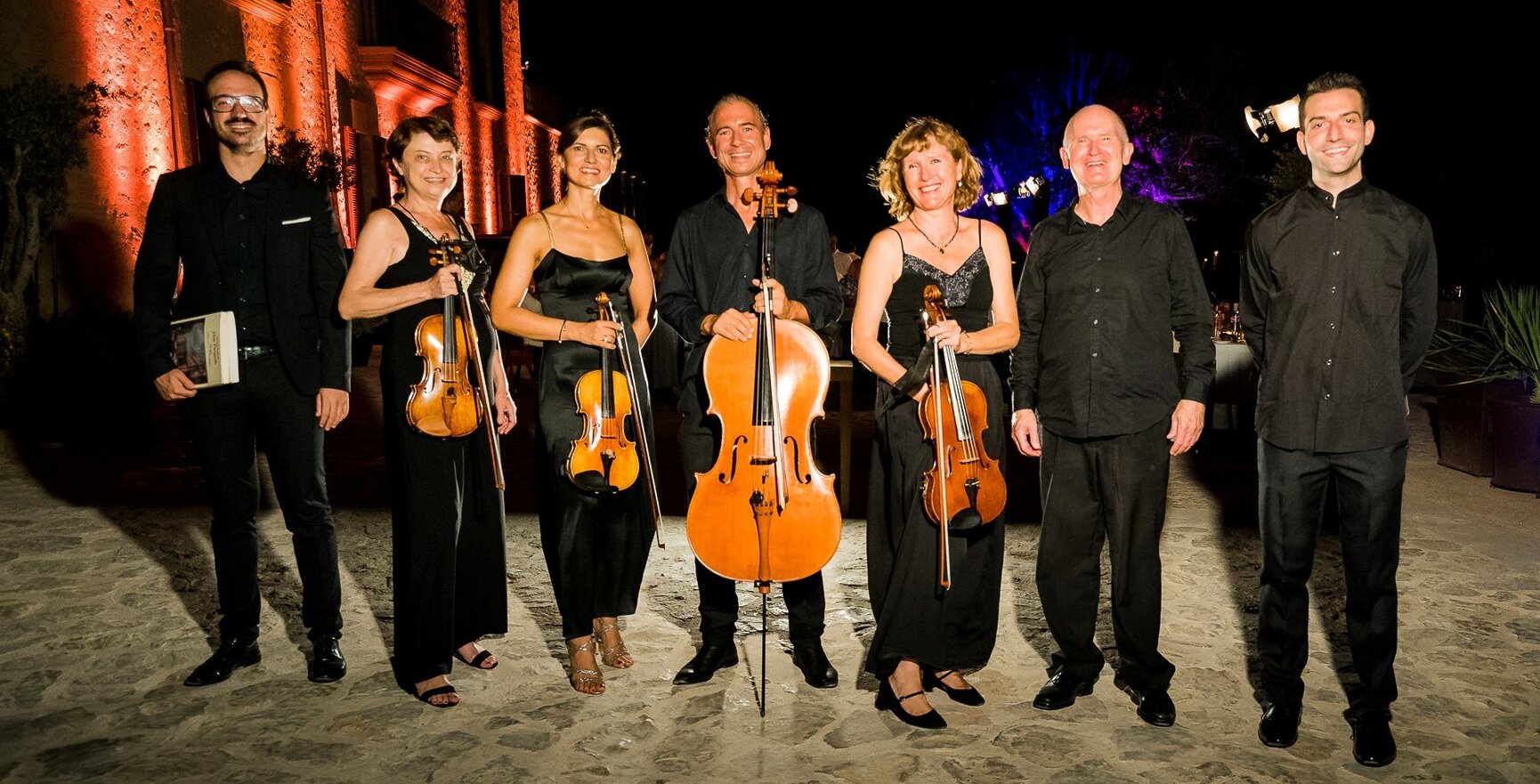 Streichquartett in schwarzer Kleidung steht auf steinerner Terrasse vor warm beleuchtetem Gebäude, bereit für Hotelabendveranstaltung
