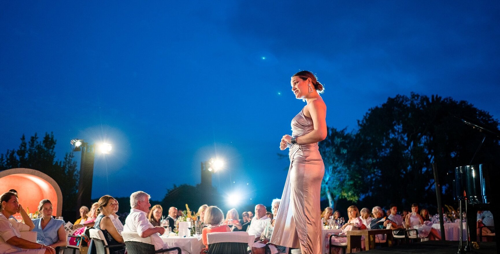 Abendliche Gala im Freien mit elegant gekleideter Frau auf der Bühne und Gästen an gedeckten Tischen unter klarem Nachthimmel
