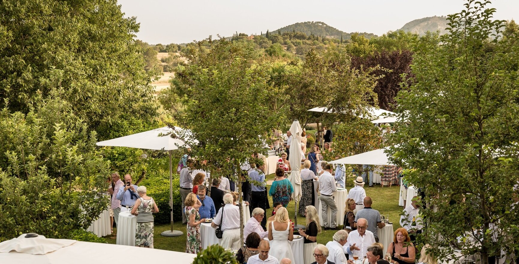 Elegante Gartenparty im Freien mit Gästen unter weißen Sonnenschirmen, umgeben von grüner Natur und entfernten Hügeln