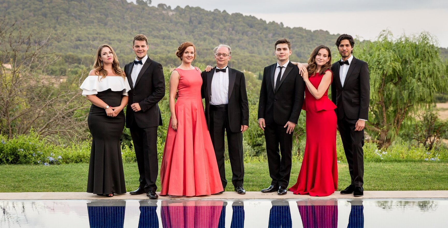 Sieben elegant gekleidete Gäste in Abendkleidern und Smokings posieren am Pool vor einer bewaldeten Hügelkulisse