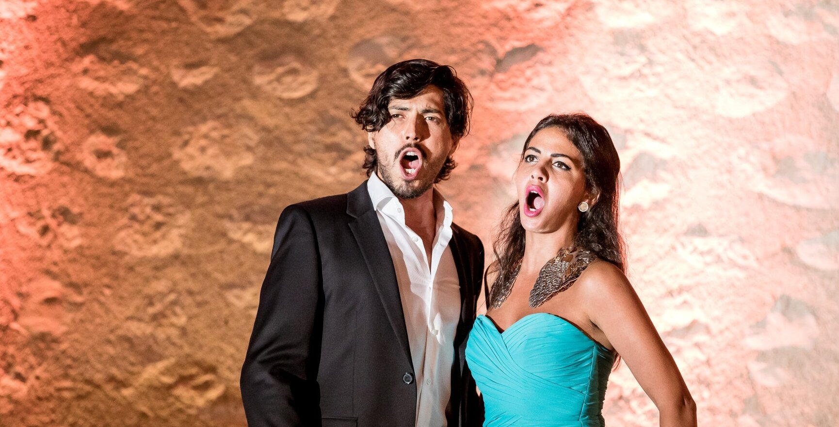 Mann im schwarzen Anzug und Frau im türkisfarbenen Kleid singen leidenschaftlich vor einer warm beleuchteten, strukturierten Wand