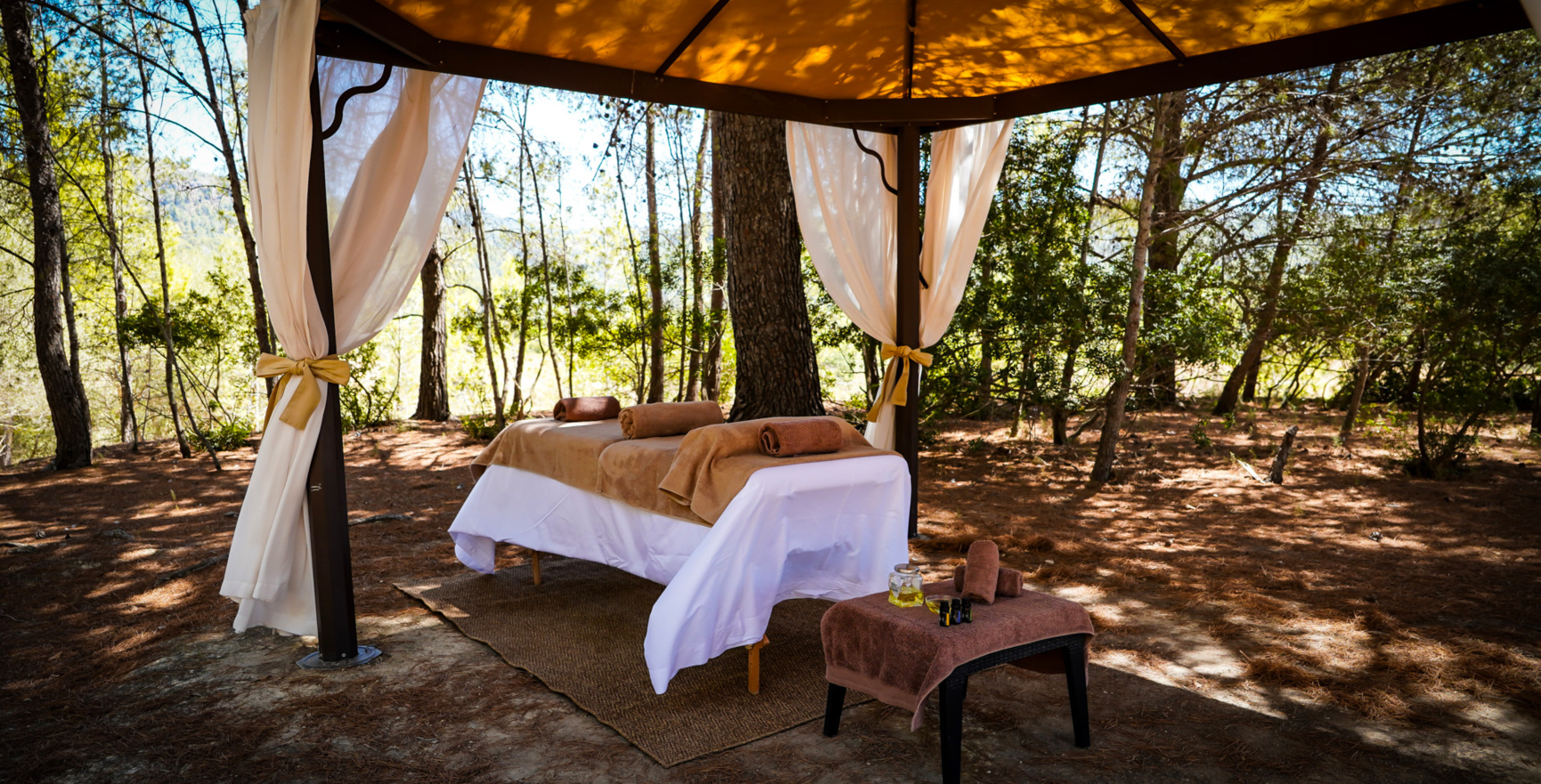 Área de masaje al aire libre bajo una carpa sombreada con cortinas suaves, toallas, aceites y rodeada de bosque de pinos tranquilo