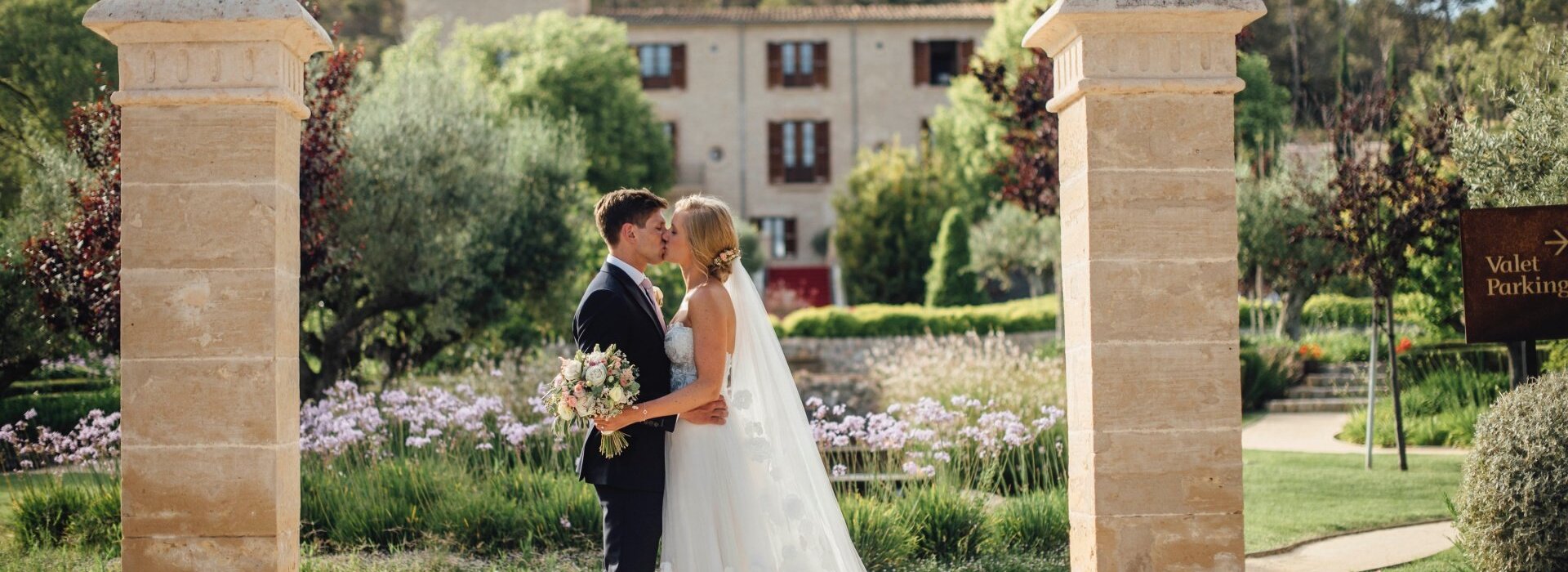 Braut und Bräutigam stehen vor dem Garten eines Luxushotels auf Mallorca und küssen sich.