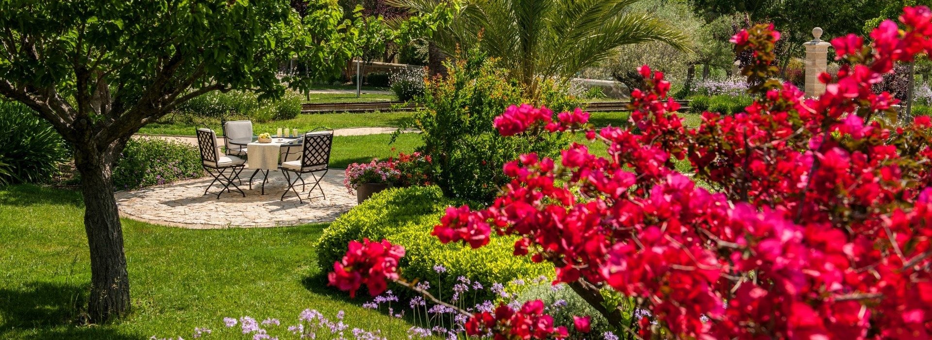 Área de comedor en jardín privado con mesa y sillas rodeada de vegetación, palmeras, flores coloridas y vistas a montañas