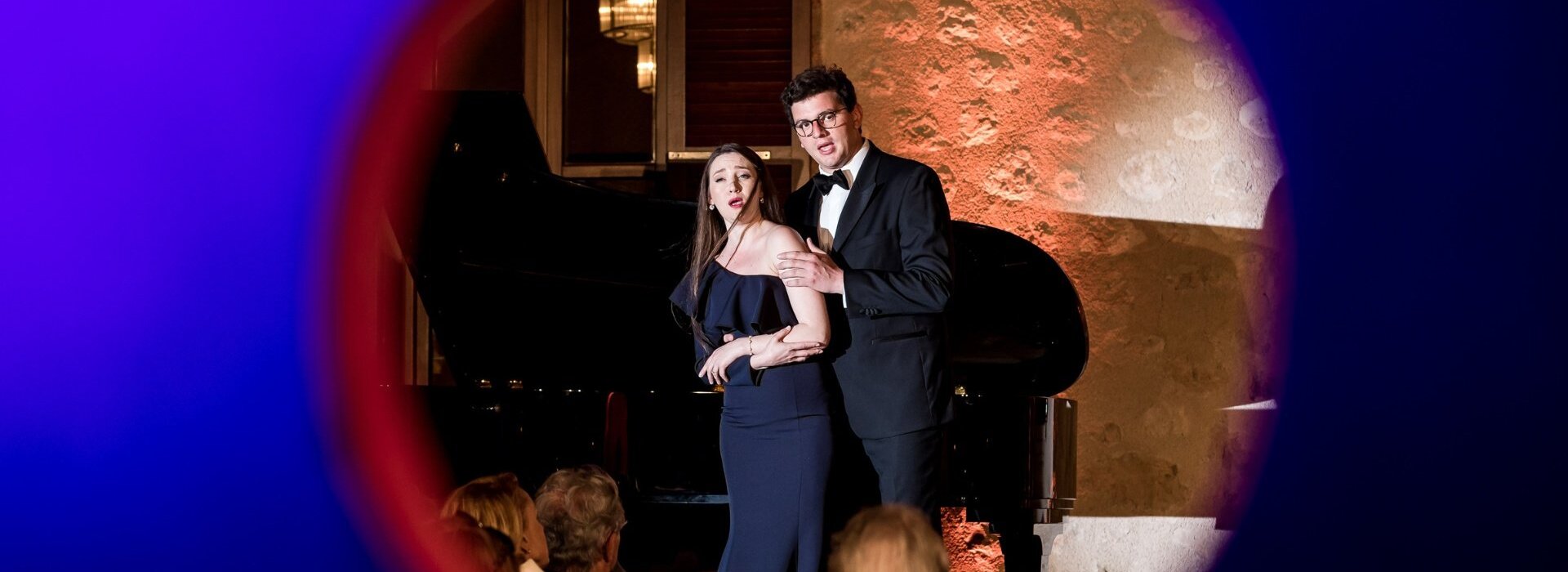 Cantantes de ópera vestidos elegantemente actuando en un lugar histórico de piedra con piano de cola y audiencia sentada