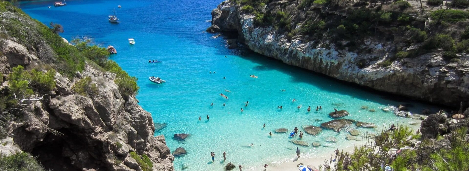 Abgelegene Bucht mit türkisfarbenem Wasser, felsigen Klippen und Gästen, die im kleinen Sandstrand mit Sonnenschirmen schwimmen und entspannen