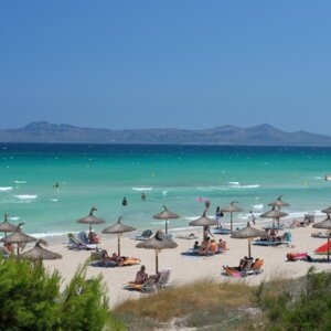 Sandstrand mit Strohdächern und Liegestühlen, Gäste sonnen sich und schwimmen im klaren türkisfarbenen Wasser mit Bergblick