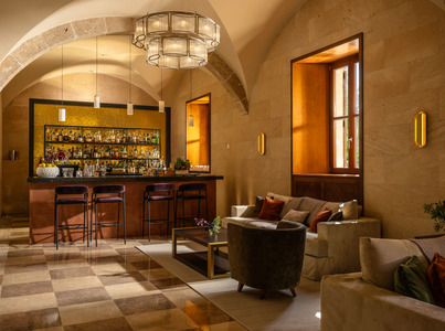 Acogedora sala de hotel con arcos de piedra, taburetes de terciopelo frente a barra con mosaicos dorados y asientos cómodos con luz natural cálida