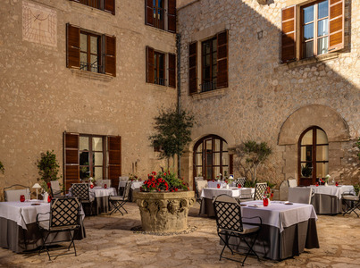 Patio exterior del hotel con muros de piedra, contraventanas de madera, mesas elegantes con manteles blancos y macetero central con flores rojas