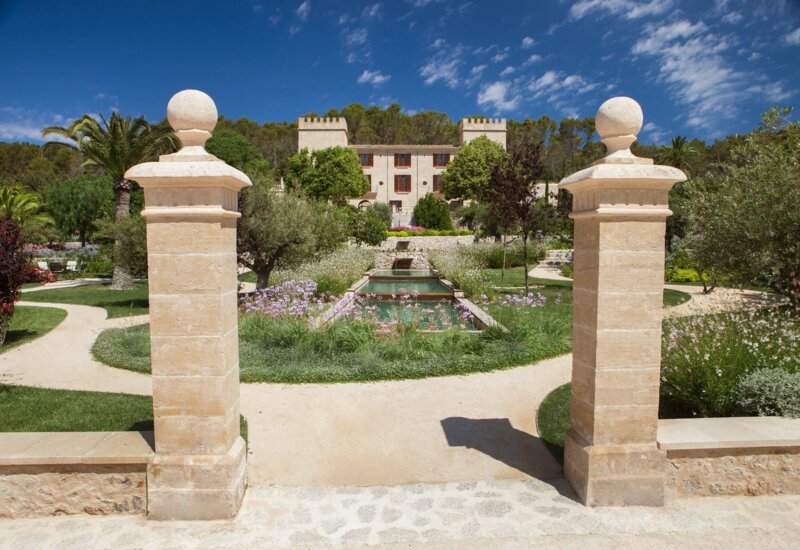 Villa histórica de piedra con jardines cuidados, estanque simétrico, palmeras y cielo azul brillante en un día soleado