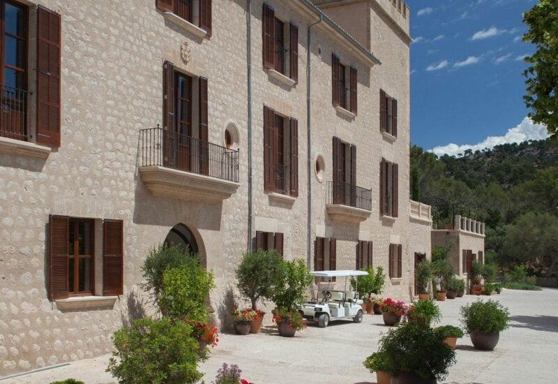 Hotel histórico de piedra con contraventanas de madera, balcones, macetas y carrito de golf en patio soleado cerca de colinas boscosas