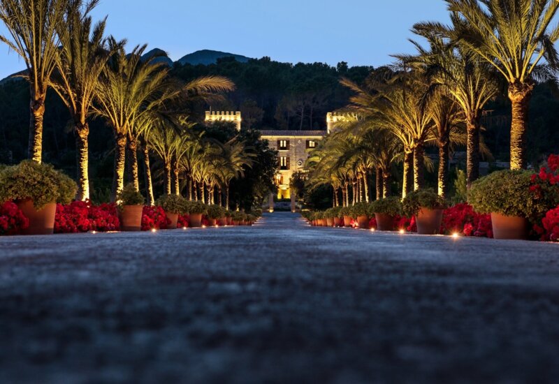 Camino bordeado de palmeras iluminadas con macetas y flores que conduce a un hotel de piedra histórico suavemente iluminado al atardecer