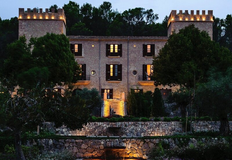 Hotel histórico de piedra con torres iluminadas, rodeado de árboles y terrazas ajardinadas al anochecer, ambiente tranquilo y elegante