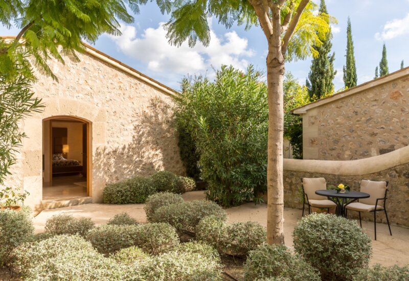 Patio acogedor de villa de piedra con vegetación abundante, área de descanso sombreada y vista a un dormitorio con luz suave