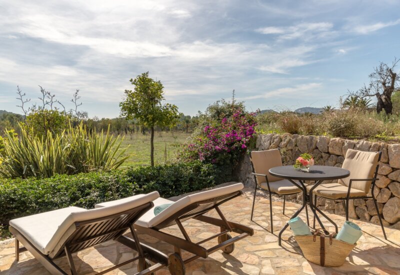 Patio soleado de piedra con tumbonas acolchadas y mesa con sillas, rodeado de abundante vegetación y flores coloridas