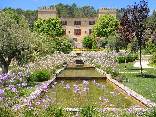 Historisches Steinhotel mit üppigen Gärten, blühenden Pflanzen und ruhigen Wasseranlagen unter klarem blauem Himmel