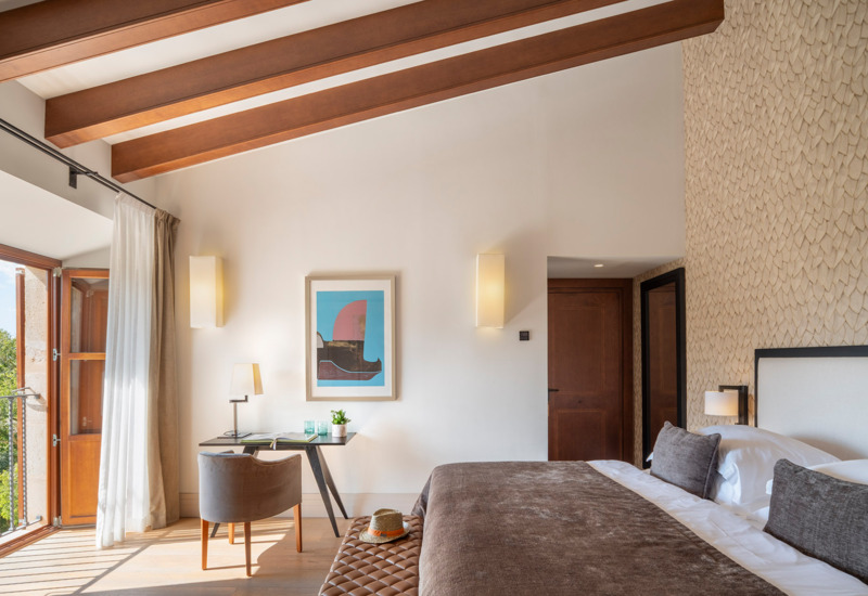 Helles Hotelzimmer mit Holzdeckenbalken, großem Bett mit brauner Bettwäsche, Schreibtisch mit Lampe und Balkontür mit Grünblick