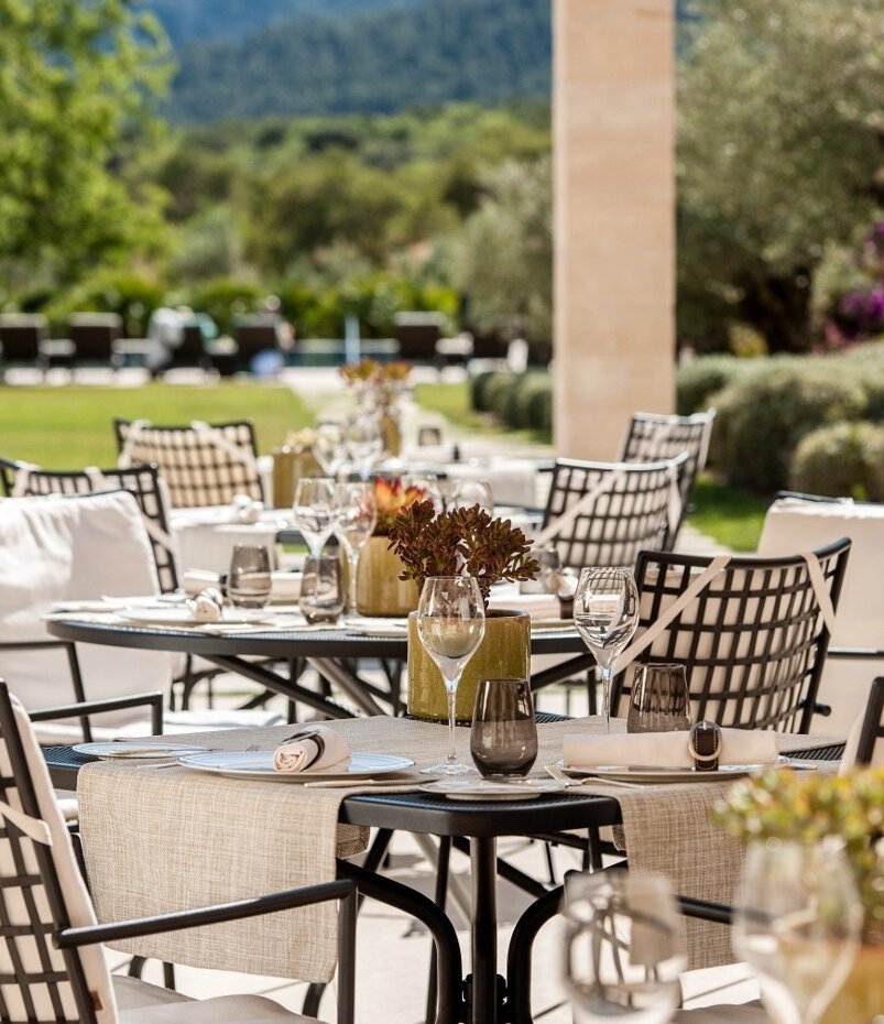 Eleganter Außenbereich mit gedeckten Tischen, gepolsterten Stühlen und Grünflächen, mit Blick auf sonnigen Garten und entfernte Hügel
