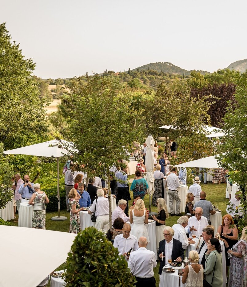 Elegante Gartenparty im Freien mit Gästen unter weißen Sonnenschirmen, umgeben von grüner Natur und entfernten Hügeln