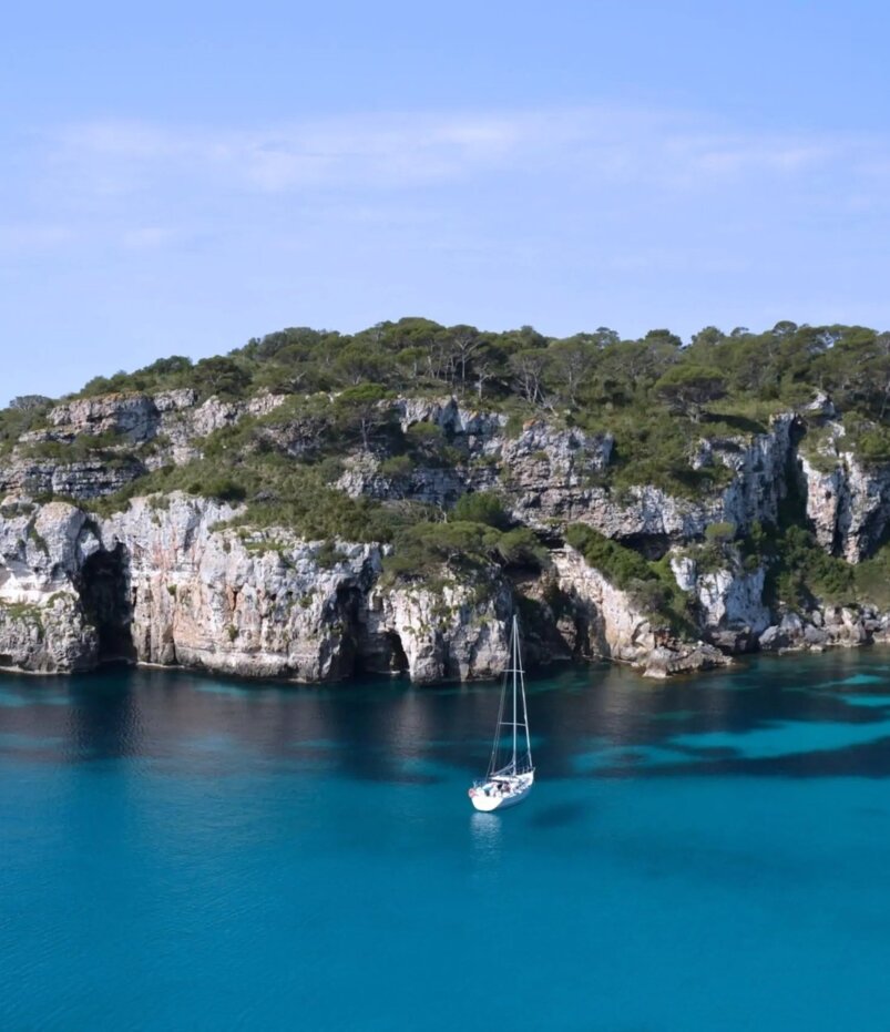 Segelboot vor felsigen, bewaldeten Klippen in klaren türkisfarbenen Gewässern mit kleinem Sandstrand.