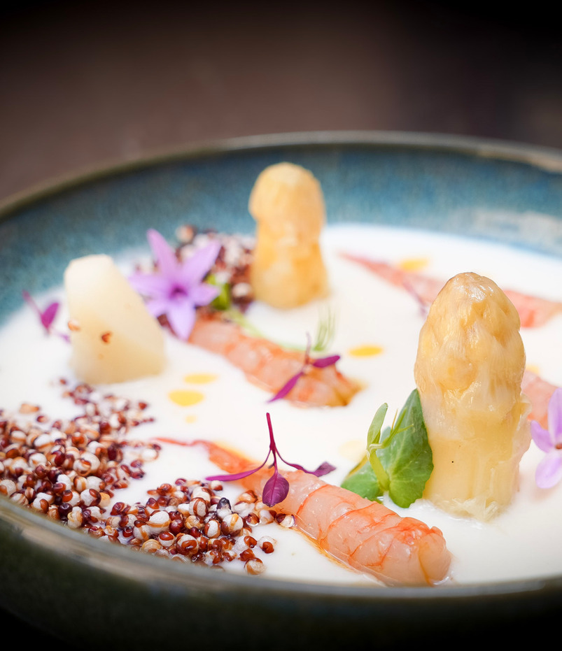 Entrante cremoso en bol azul: langostinos, espárragos blancos, quinoa roja, flores moradas comestibles y microverdes.
