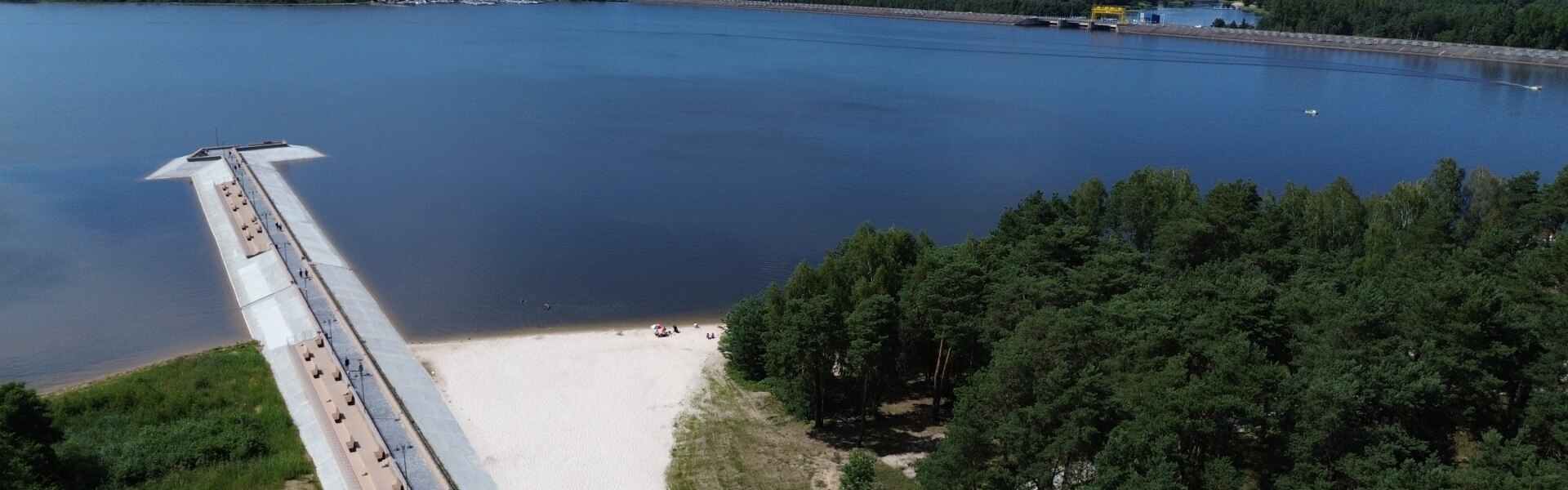 Piaskowa plaża nad jeziorem z długim pomostem spacerowym, ławkami i sosnowym lasem — idealna na relaks, spacery i kąpiele