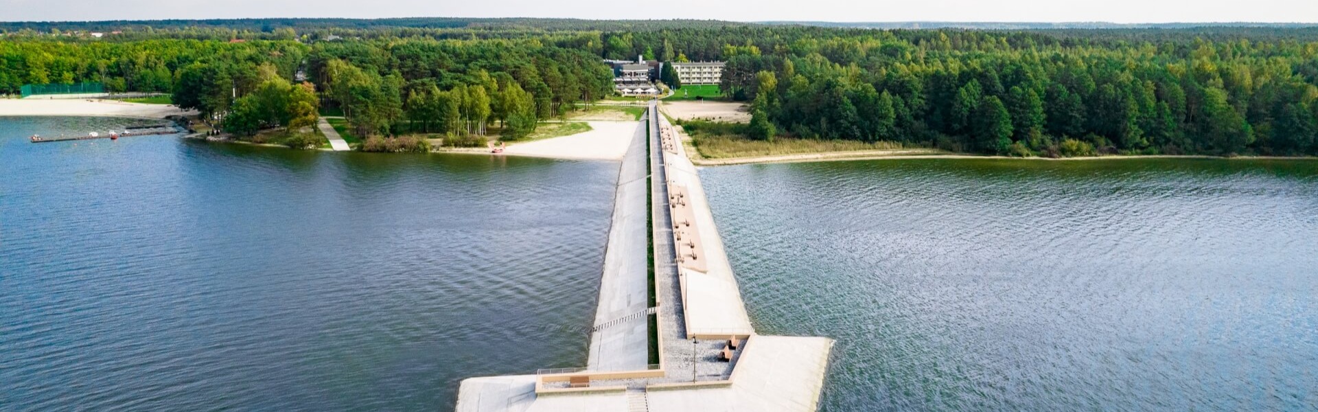 Długie betonowe molo do jeziora z piaszczystą plażą, nadbrzeżnym ośrodkiem z tarasami, otoczone sosnowym lasem i spokojną wodą.