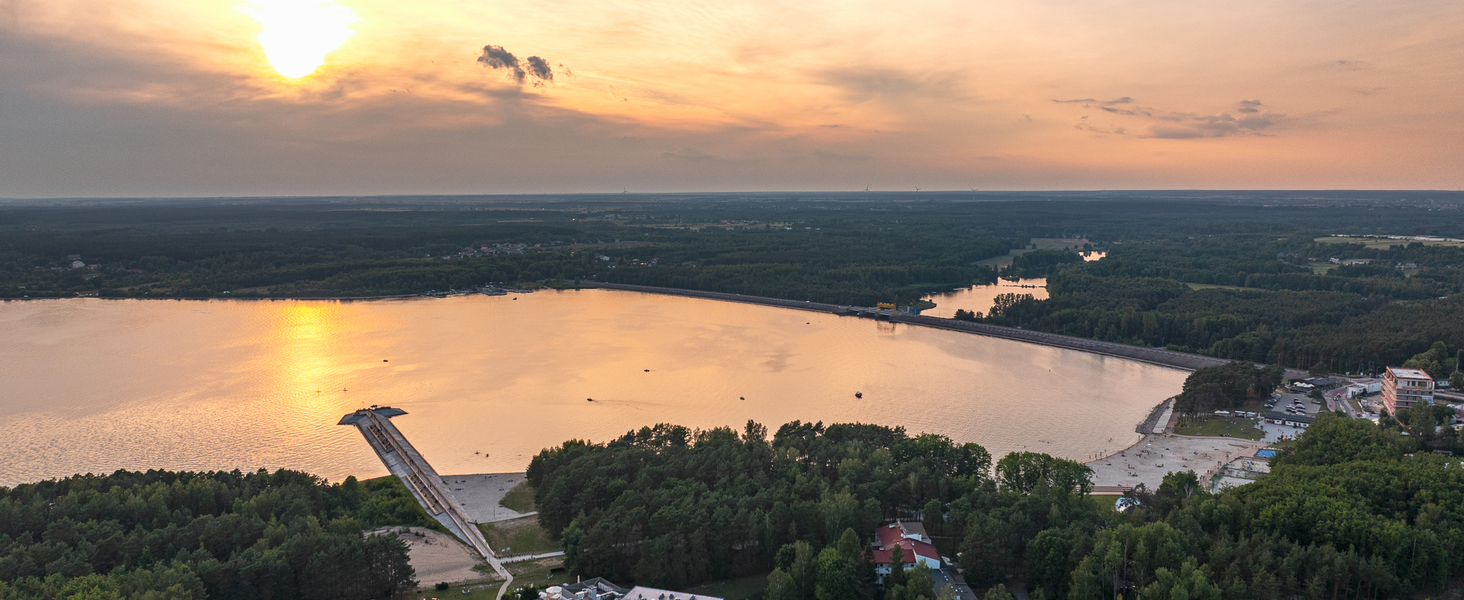 Zachód słońca nad jeziorem z długim molo, piaszczystą plażą, otaczającym lasem i kompleksem hotelowym przy brzegu.