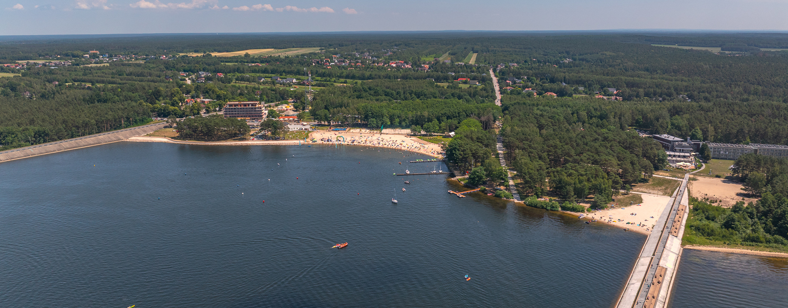 Kurort nad jeziorem z piaszczystą plażą, molem i hotelem przy lesie; kąpieliska, kajaki i leżaki na plaży.