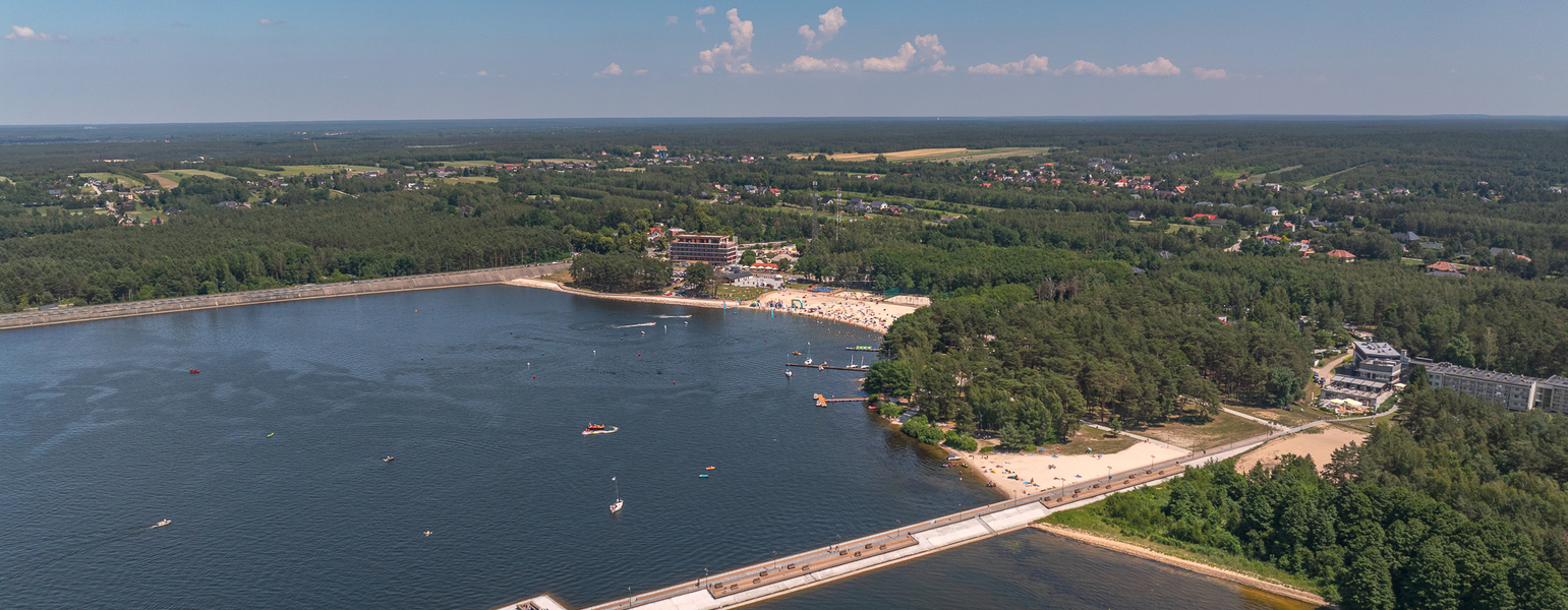 Kurort nad jeziorem z piaszczystą plażą, molo, hotelem przy lesie i wodnymi atrakcjami (łodzie, kąpieliska)