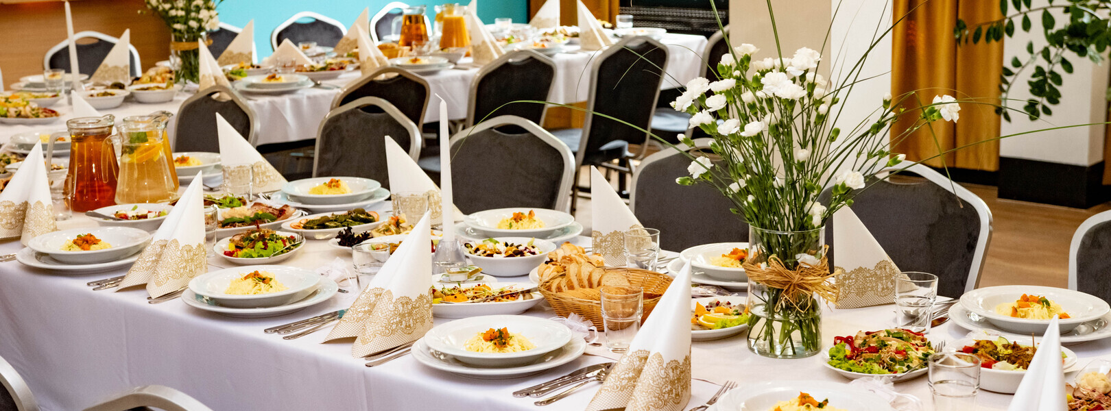 Sala bankietowa z długim stołem w białych obrusach, eleganckimi serwetkami, talerzami z makaronem, sałatkami i bukietami goździków.