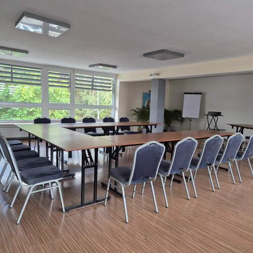 Sala konferencyjna z ustawionymi w podkowę stołami, szarymi krzesłami, flipchartem, projektorem i dużymi oknami z widokiem na zieleń.