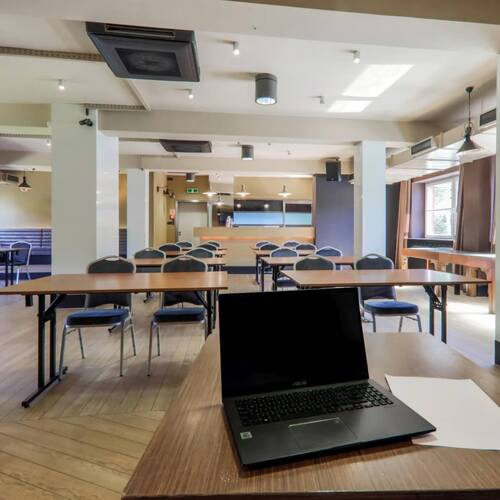 Sala szkoleniowa z rzędami stolików i krzeseł, drewniana podłoga, duże okna i bar z tyłu, laptop na stole.