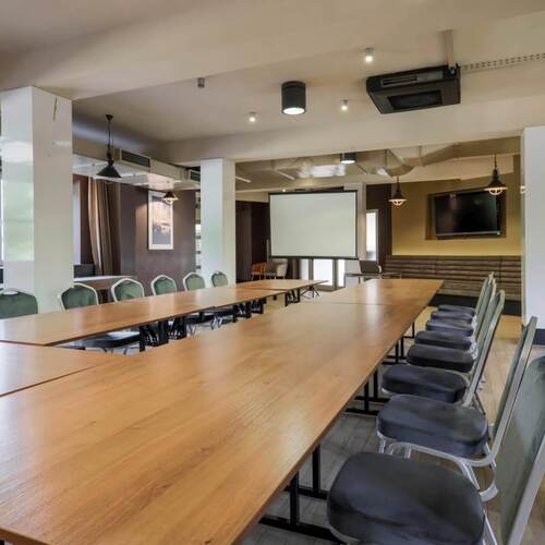 Sala szkoleniowa z U‑kształtnym stołem drewnianym, krzesłami, ekranem projekcyjnym, telewizorem i wiszącymi lampami.