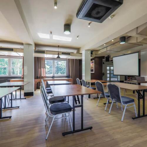 Sala konferencyjna z ustawionymi stołami i krzesłami, ekranem projekcyjnym, dużymi oknami i drewnianą podłogą.