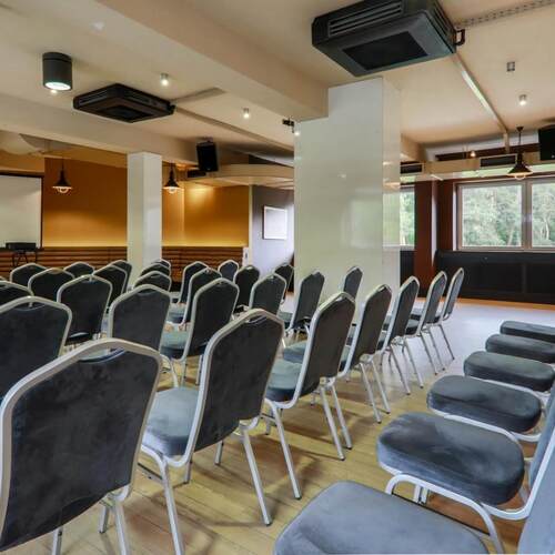 Sala konferencyjna w układzie teatralnym z szarymi krzesłami, ekranem projekcyjnym, nagłośnieniem, drewnianą podłogą i oknami z widokiem na las