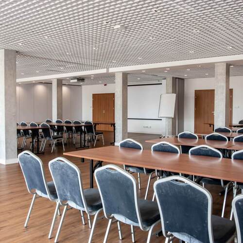 Przestronna sala konferencyjna z ustawionymi stołami, wygodnymi krzesłami, ekranami i flipchartem.