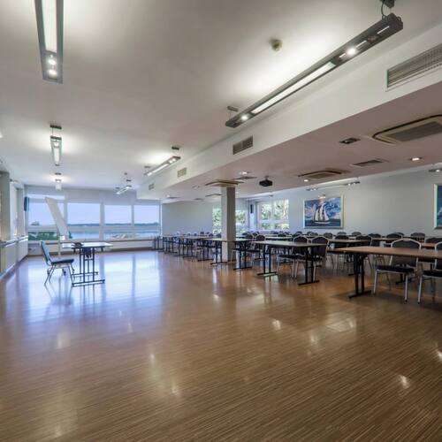 Sala konferencyjna z ustawionymi stołami i krzesłami, ekranem projekcyjnym i flipchartem, nowoczesnym oświetleniem i dużymi oknami na jeziorę.
