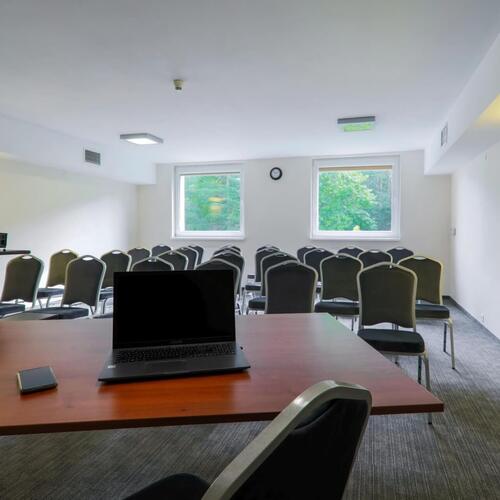Sala konferencyjna z rzędami krzeseł, stołem z laptopem i projektorem, flipchart, dwa okna z widokiem na zieleń — jasne, szkoleniowe wnętrze.