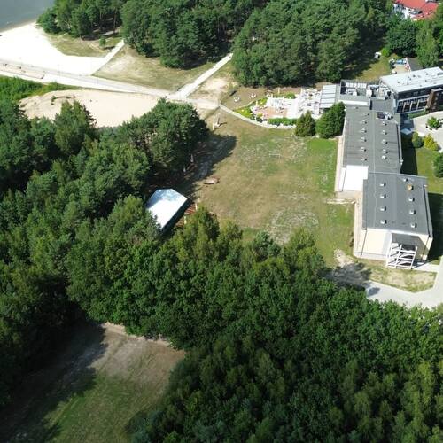 Hotel przy lesie i piaszczystej plaży z tarasem zewnętrznym, ścieżkami spacerowymi, parkingiem i pomostem nad jeziorem.