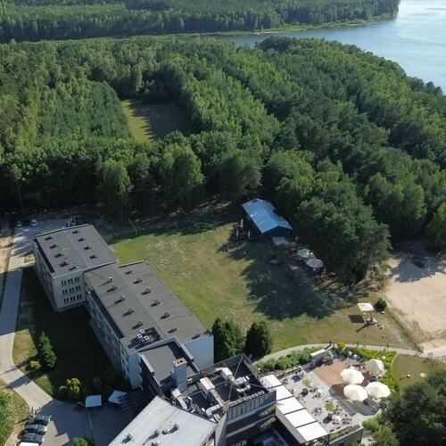 Hotelowy kompleks przy jeziorze: budynek z tarasem i parasolami, piaszczysta plaża, ścieżki i gęsty las