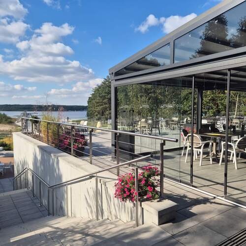 Szklany taras restauracyjny z krzesłami i stolikami, nowoczesna bryła, donice z różowymi kwiatami, widok na plażę i jezioro.