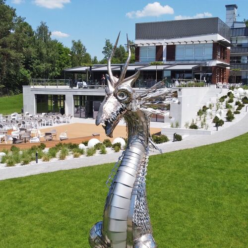Nowoczesny hotel z przestronnym tarasem i ogródkową restauracją, zadbane trawniki i nasadzenia, efektowna metalowa rzeźba smoka na pierwszym planie.