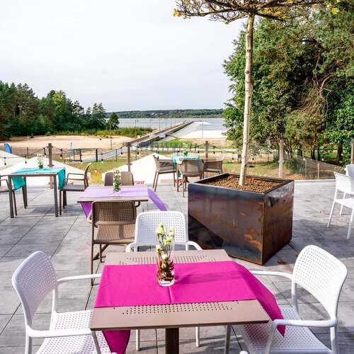 Taras restauracyjny z udekorowanymi stolikami, białymi krzesłami, donicą z drzewkiem i widokiem na plażę, molo i jezioro.