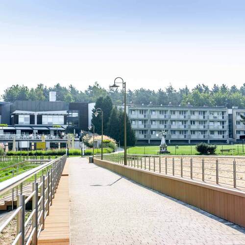 Promenada z barierką prowadząca do hotelu z tarasem na świeżym powietrzu, stolikami, balkonami pokoi, latarniami i zielonym trawnikiem