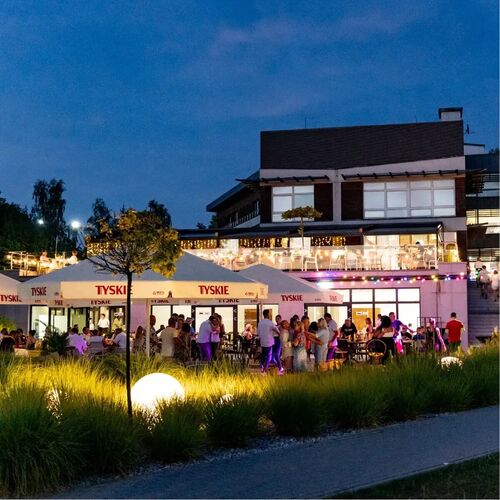 Wieczorny taras restauracyjny przy hotelu z parasolami TYSKIE, girlandami świateł, podświetlonymi kulami w trawie i bawiącymi się gośćmi.