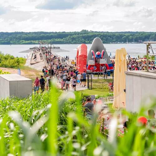 Ruchliwy nadbrzeżny festyn: zatłoczone molo i piaszczysta plaża, duża dmuchana zjeżdżalnia, taras i leżaki przy wodzie