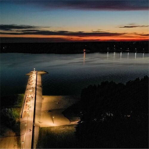 Wieczorny widok z hotelowego balkonu na oświetlony pomost, piaszczystą plażę i spokojne jezioro w barwach zachodu słońca.