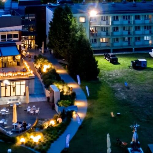 Hotelowy budynek nocą z przytulnie oświetloną tarasową restauracją, stolikami na patio, balkonami i trawnikiem